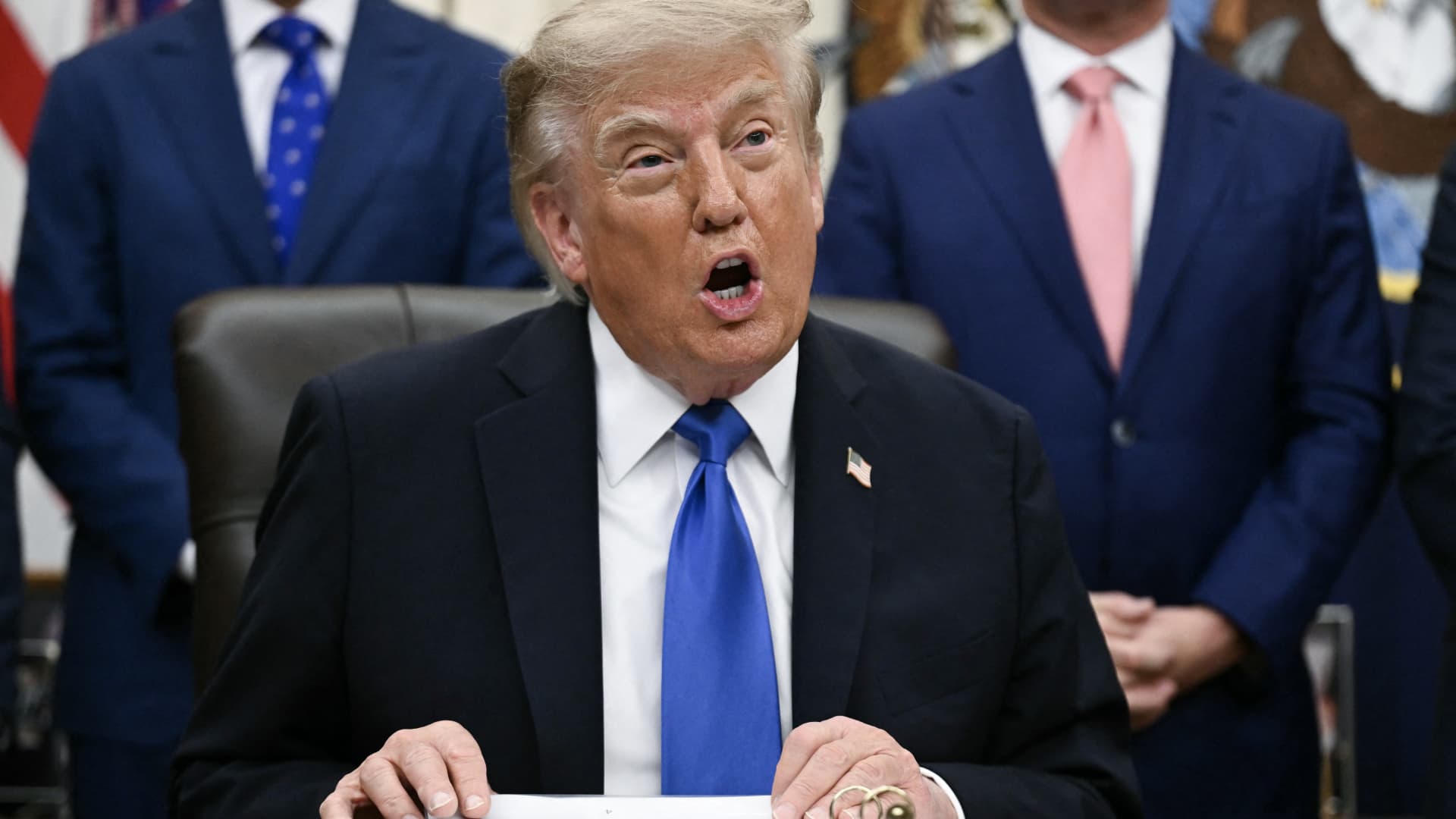 Donald Trump speaks while seated at a desk, wearing a dark suit and blue tie.