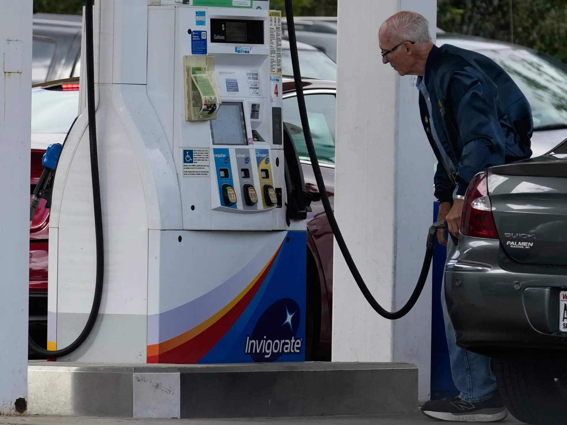 An older man refuels a gray car beside a gas pump at a station.