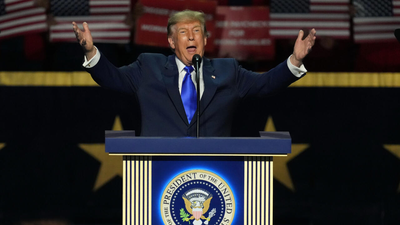 Donald Trump speaks at a podium with arms raised before an American flag backdrop.