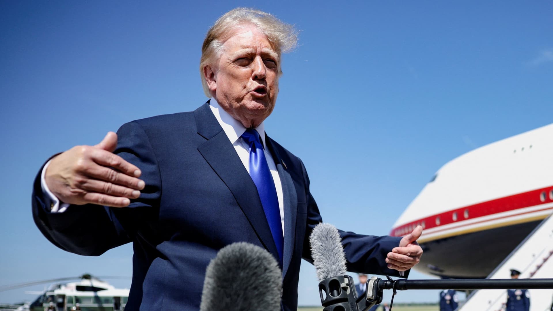 Donald Trump gestures while speaking to reporters beside microphones, with an airplane visible behind him.
