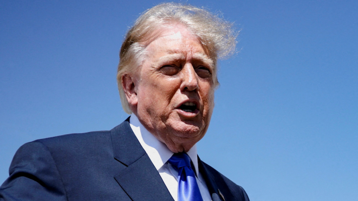 Donald Trump speaks outdoors in a dark suit and blue tie beneath a clear blue sky.