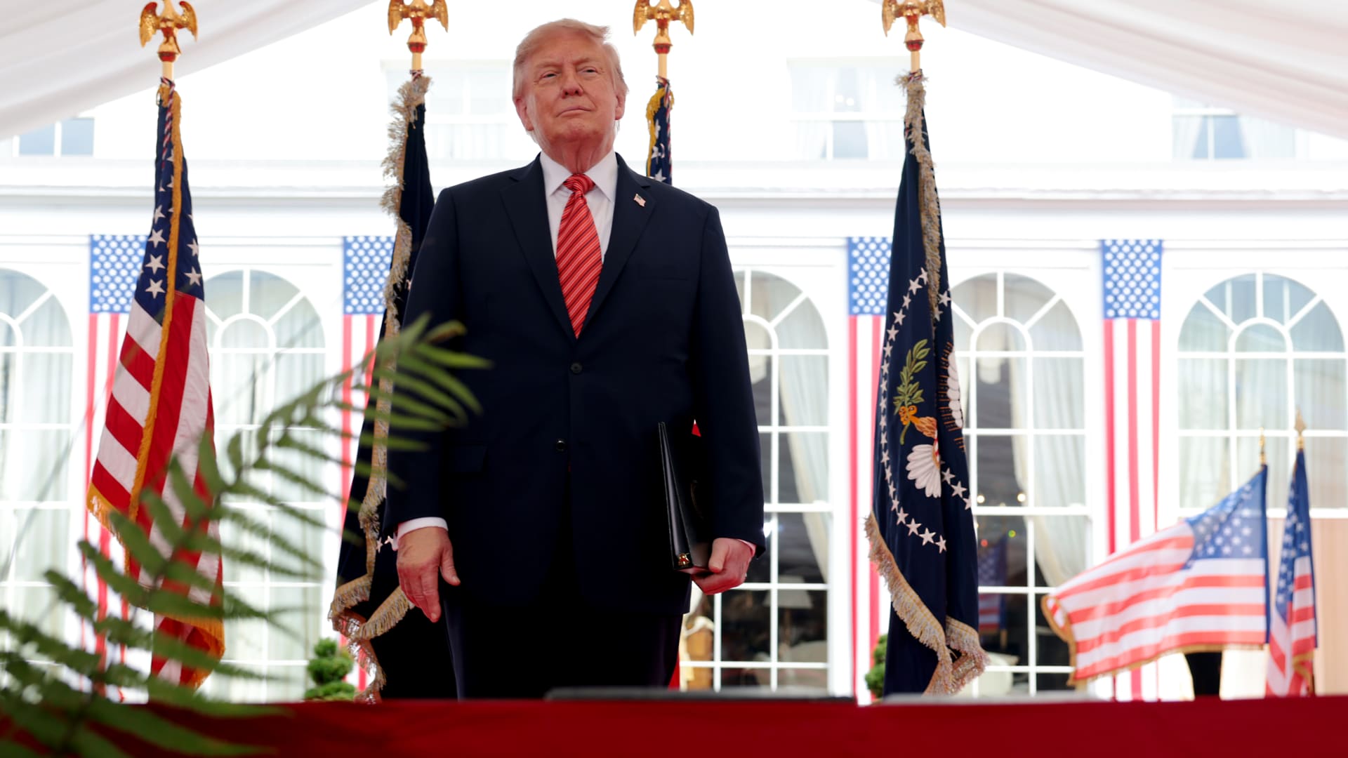 Donald Trump stands in a dark suit before American flags and presidential banners.