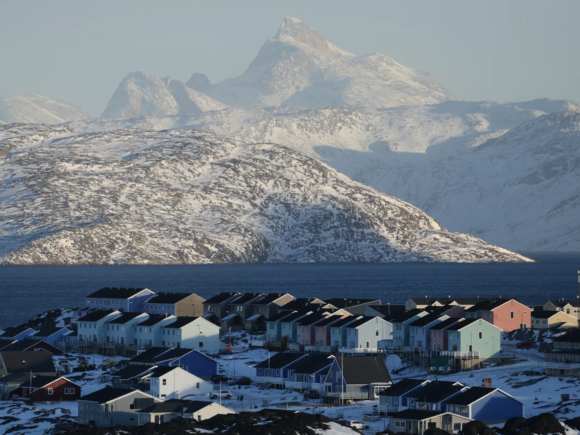 Snow-covered coastal town beneath rugged mountains, with dark blue water in the foreground.