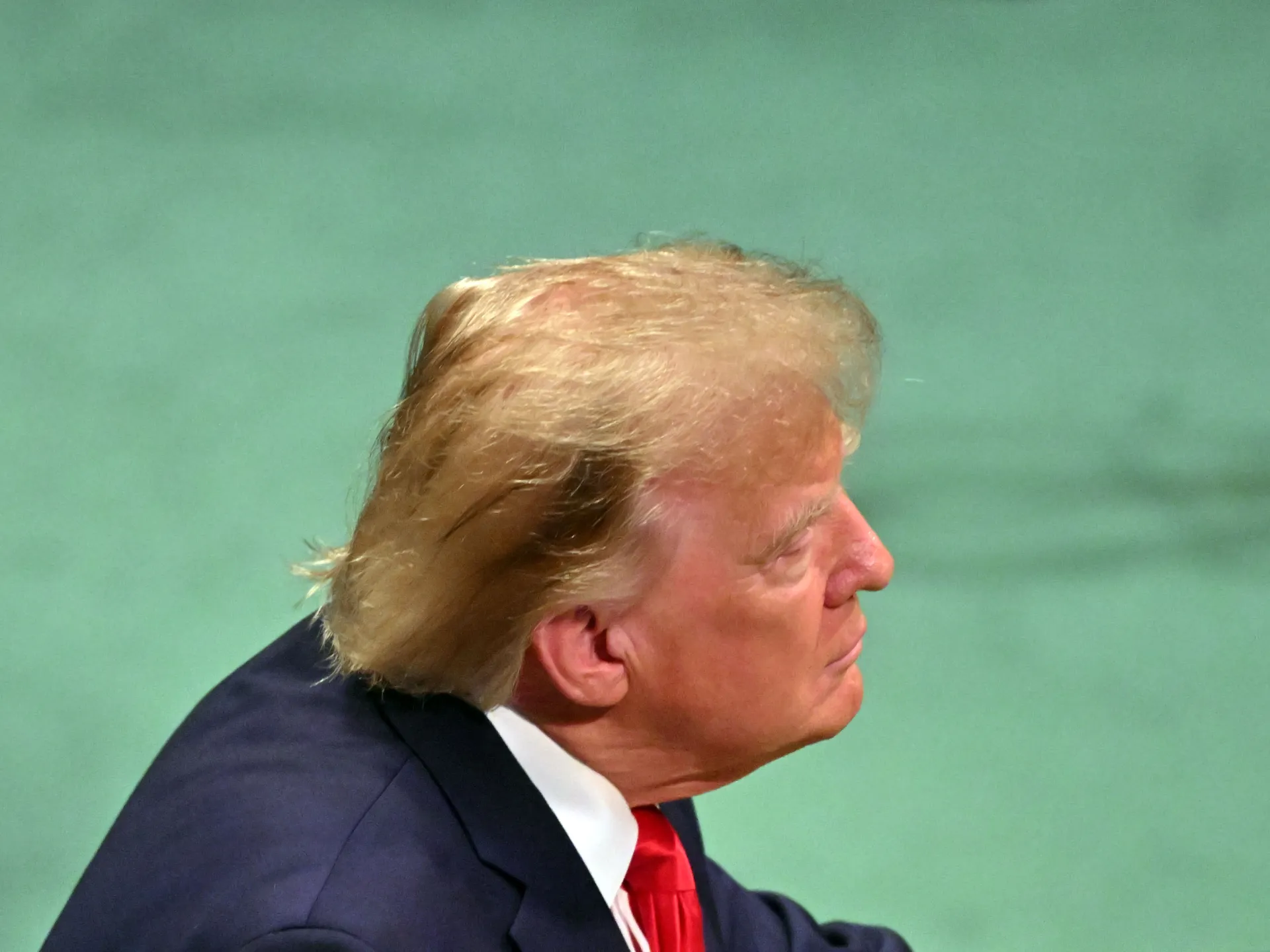 Profile view of a blond-haired man in a dark suit and red tie against a green background