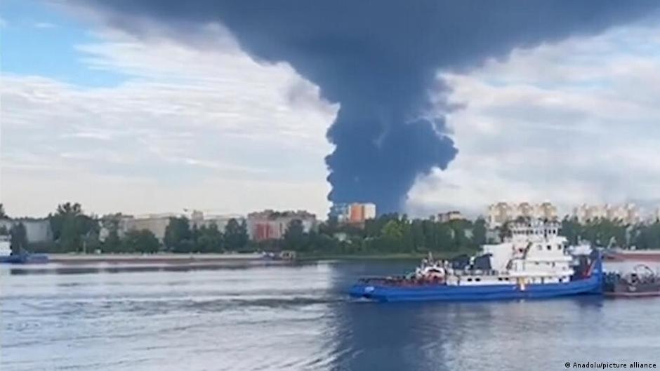 Blue-and-white tugboat crosses water before a large dark smoke plume rising over a shoreline.