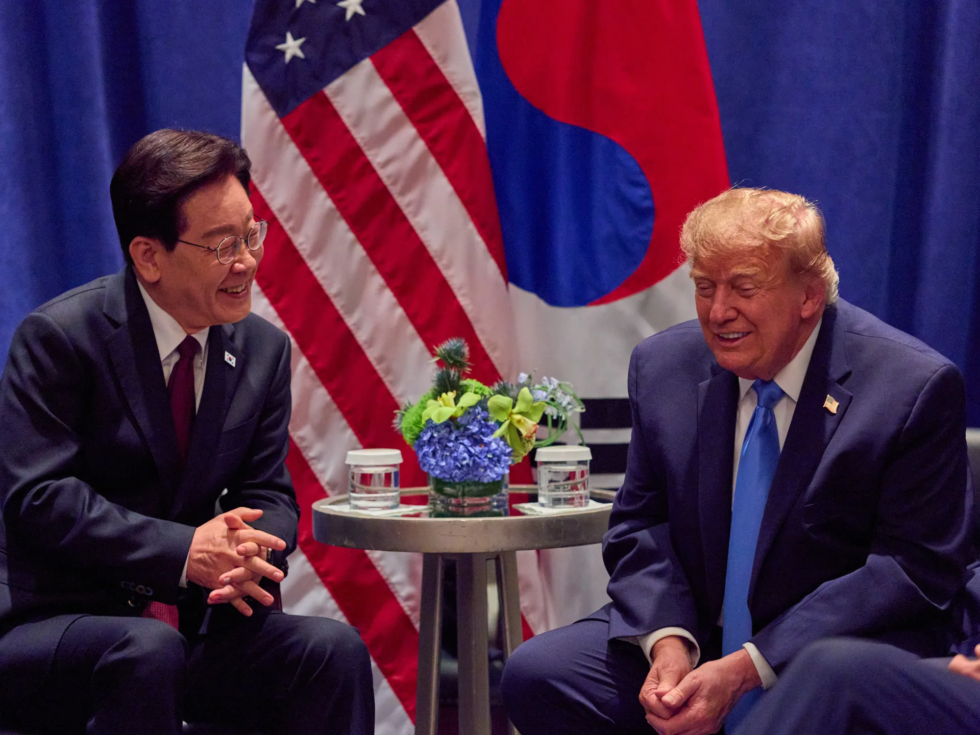 Two suited men sit facing each other beside a flower arrangement, with U.S. and South Korean flags behind them.