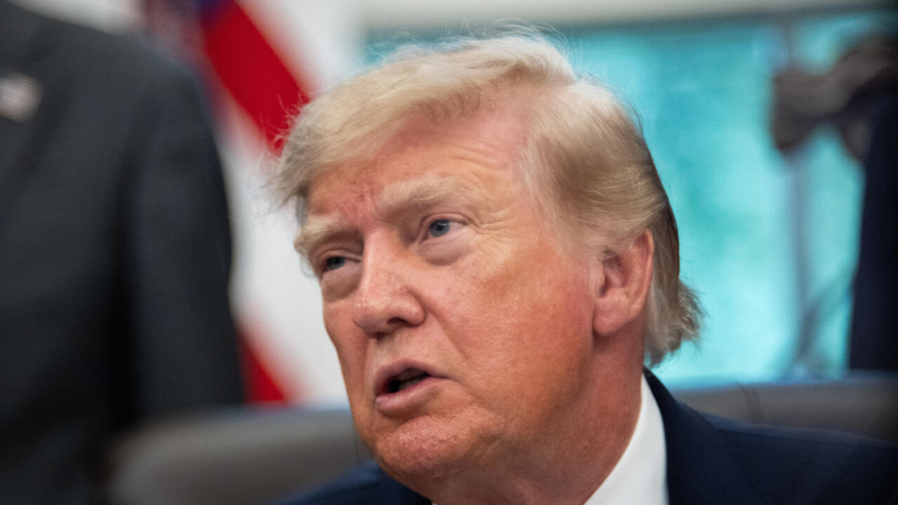 Close-up of Donald Trump speaking, with a blurred U.S. flag and people in the background.