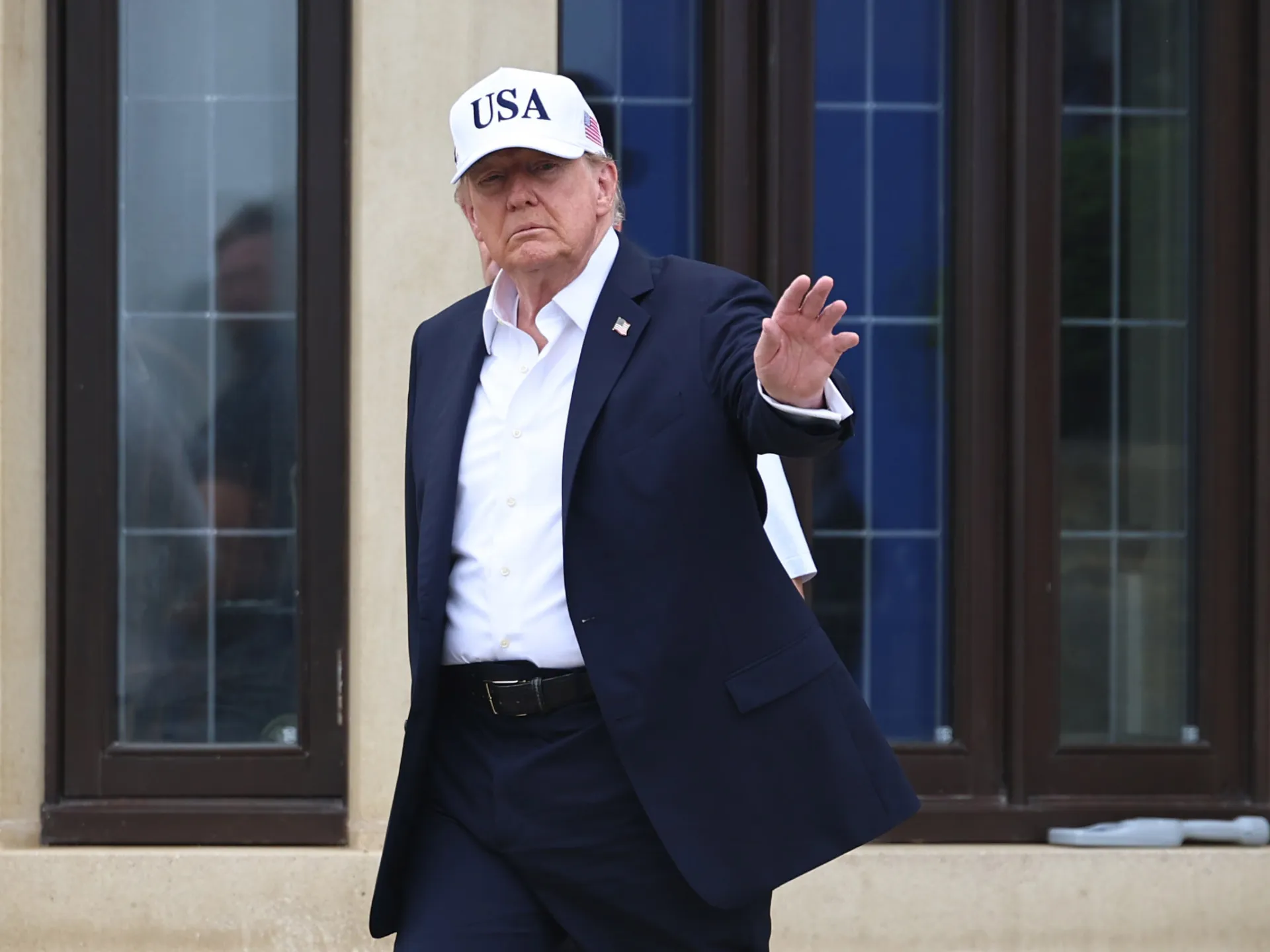 Donald Trump in a dark suit and USA cap gestures outside a building.