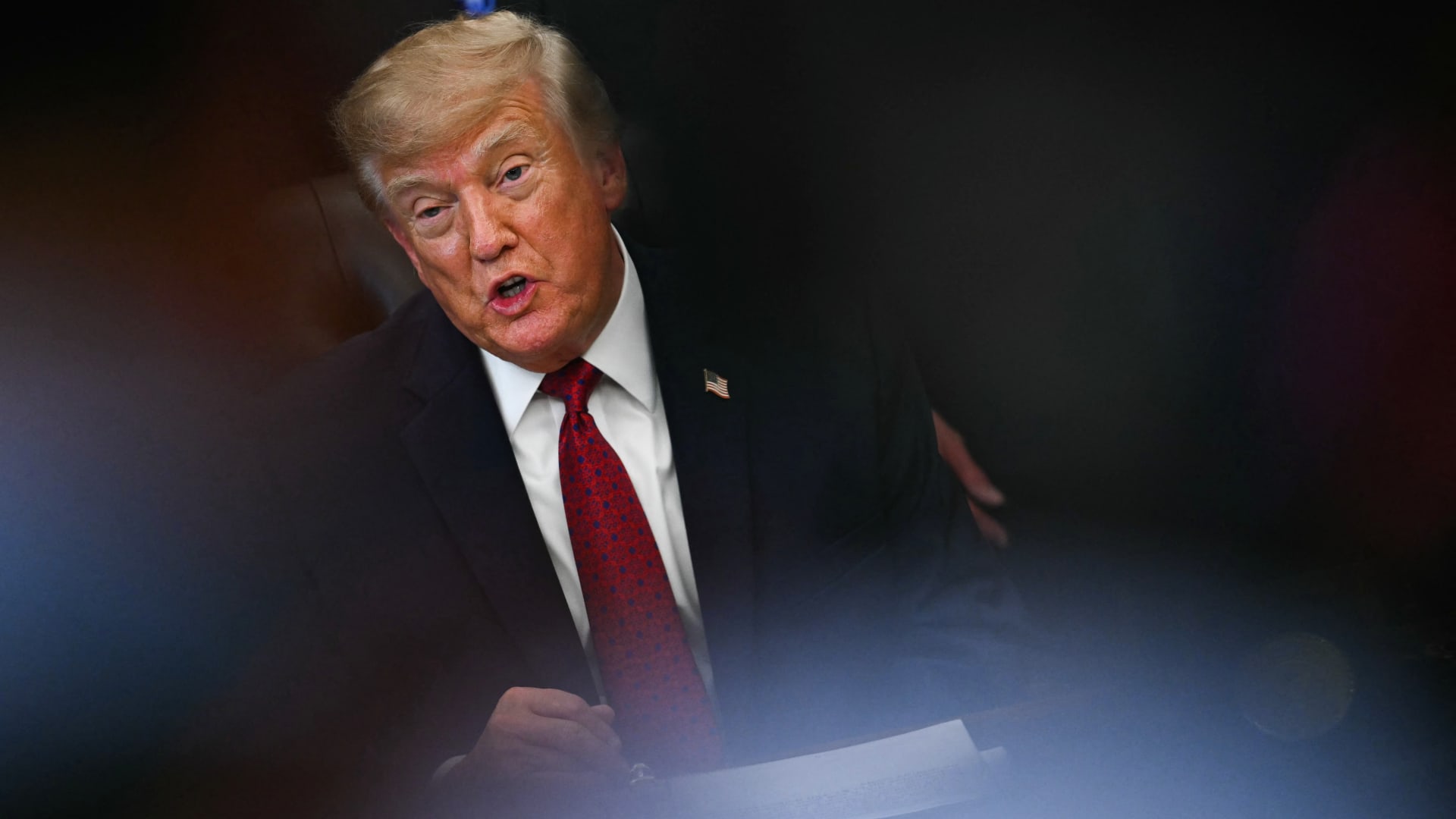 Donald Trump in a dark suit and red tie, speaking behind a partially obscured foreground.