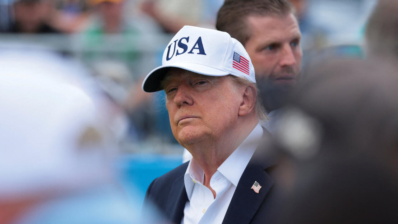 Donald Trump wearing a USA cap and suit amid a blurred crowd.