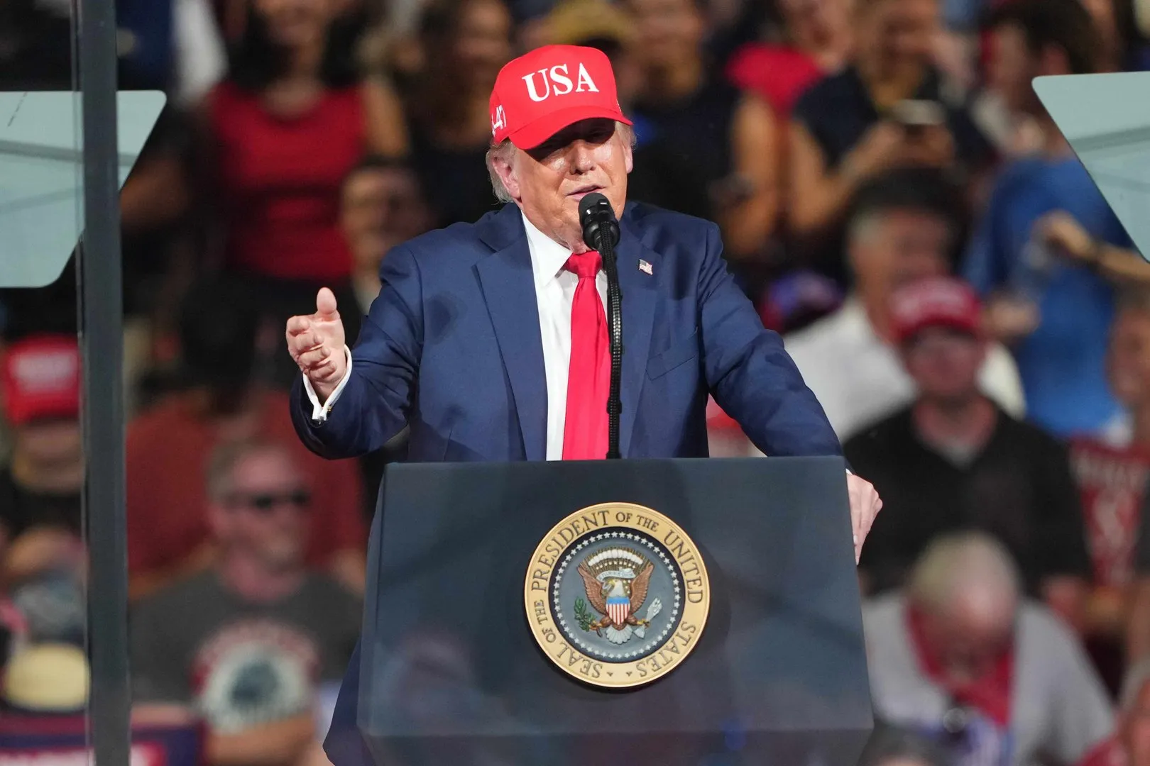 Donald Trump speaks at a podium wearing a red USA cap before a blurred crowd.