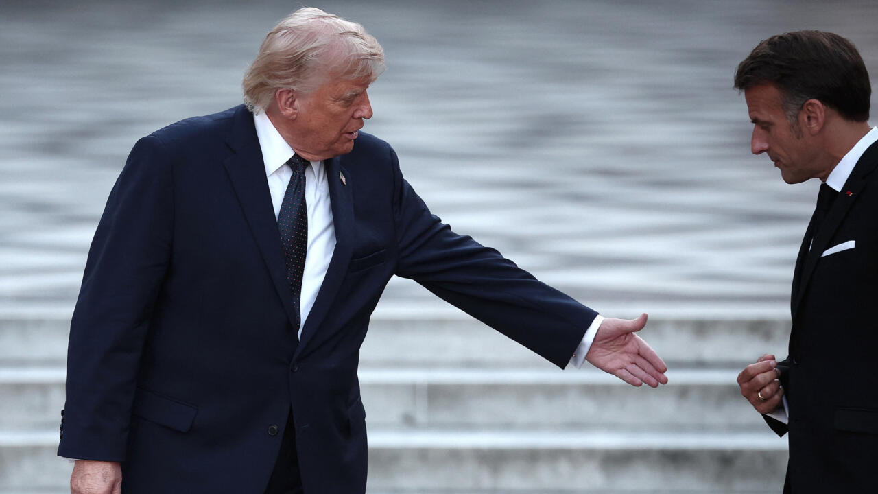 Two suited men stand beside water, one extending his hand toward the other.