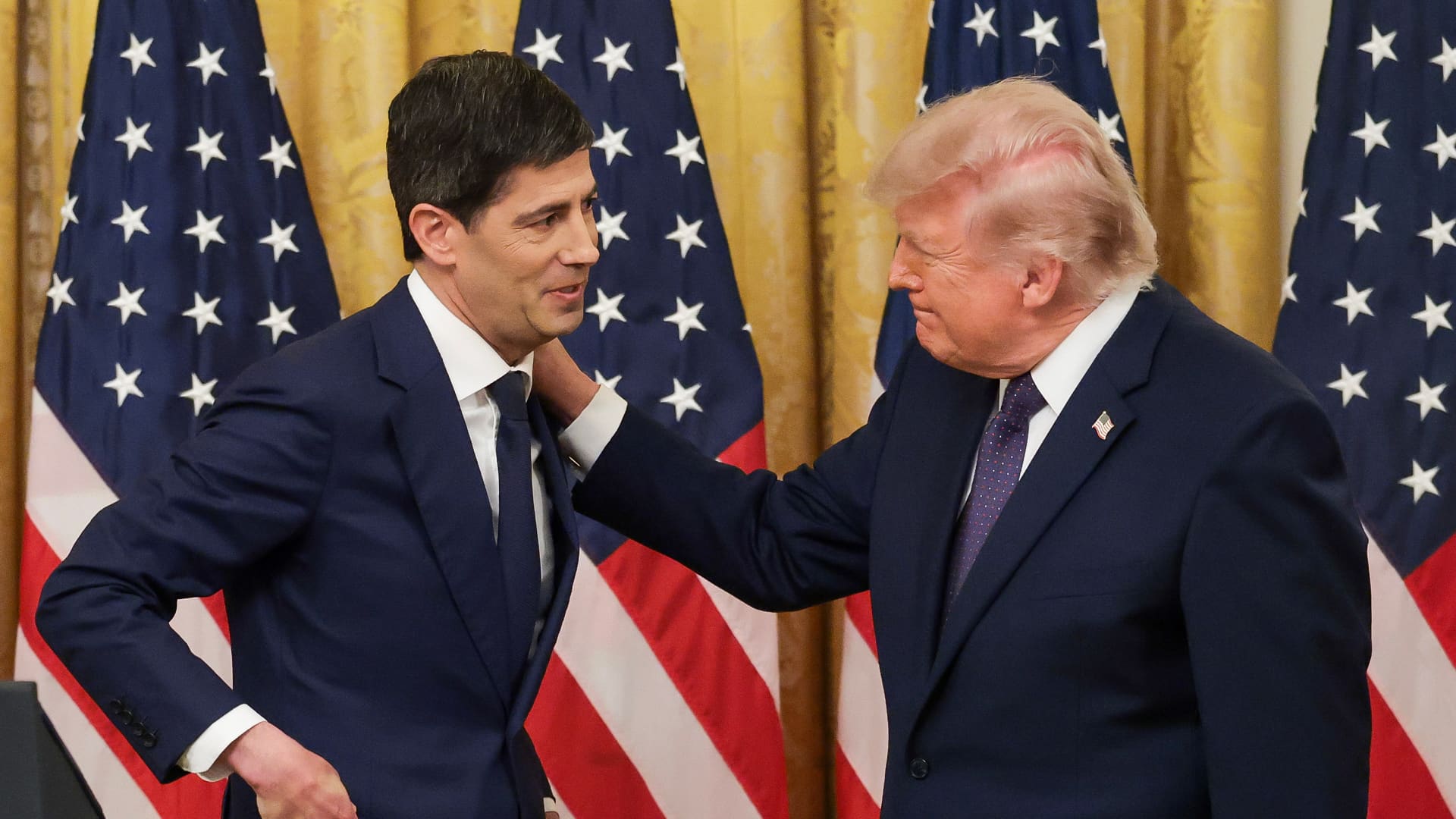 Donald Trump gestures toward a suited man as American flags hang behind them