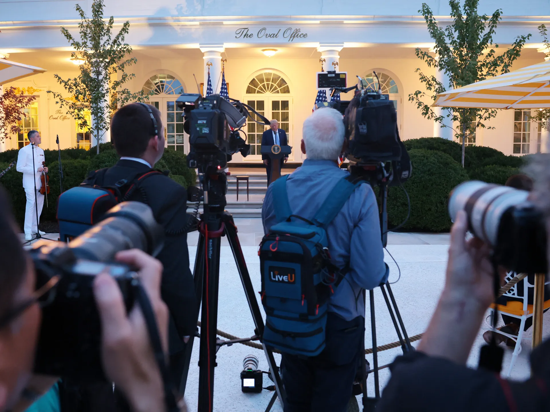 Television cameras and photographers film a speaker at a podium outside a columned building.