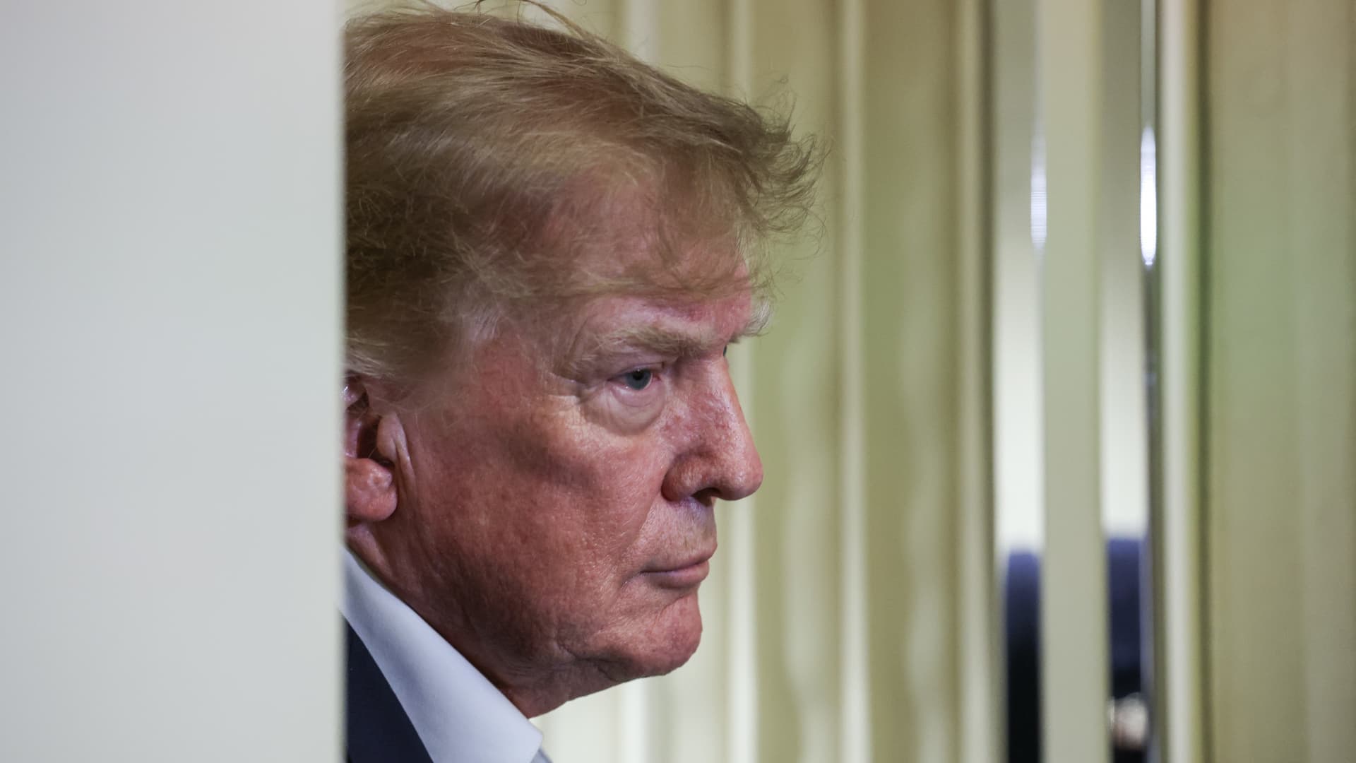 Donald Trump shown in side profile beside pale curtains, looking toward the right side of the frame.