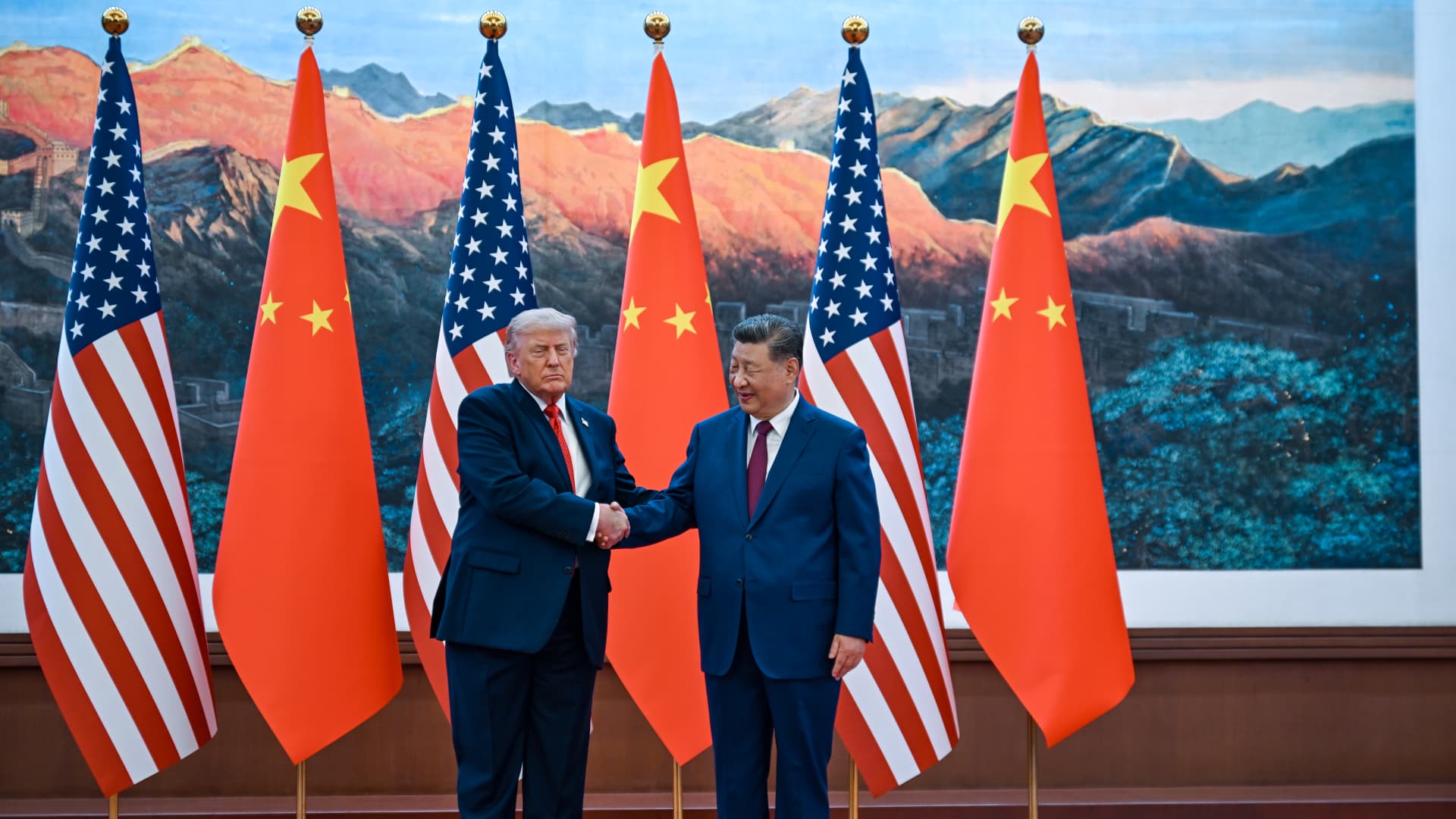 Two suited leaders shake hands before alternating American and Chinese flags and a mountain landscape.