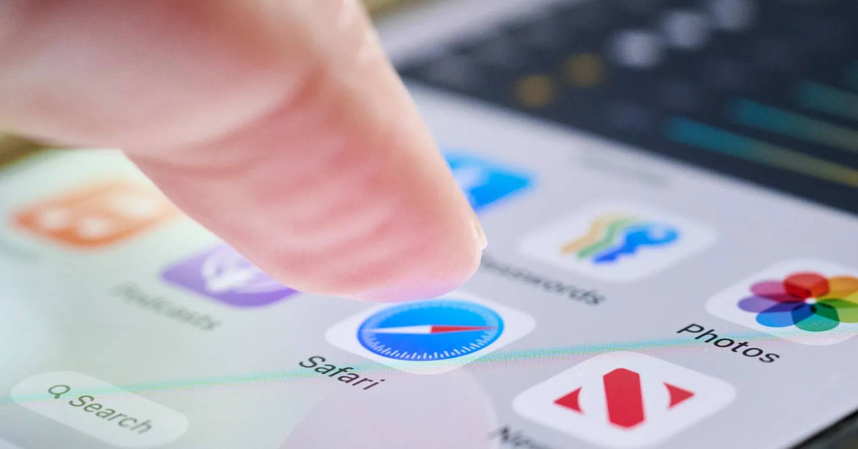 Finger tapping an iPhone home screen with Safari, Photos, and other app icons visible.