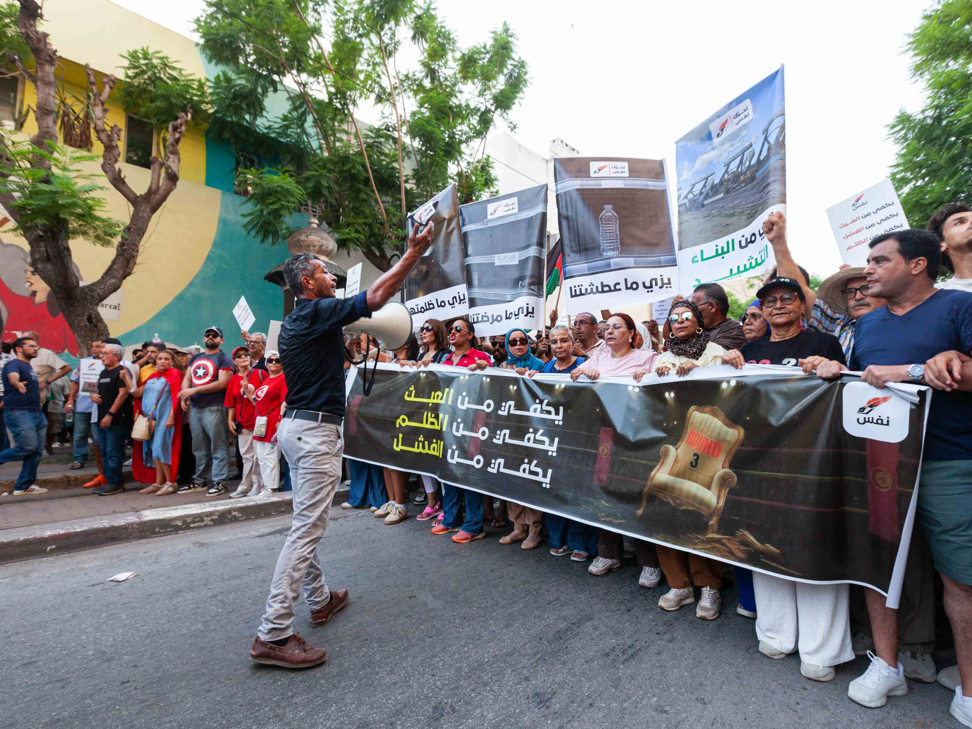 A street crowd holds a large black banner and signs, while a man raises a megaphone.