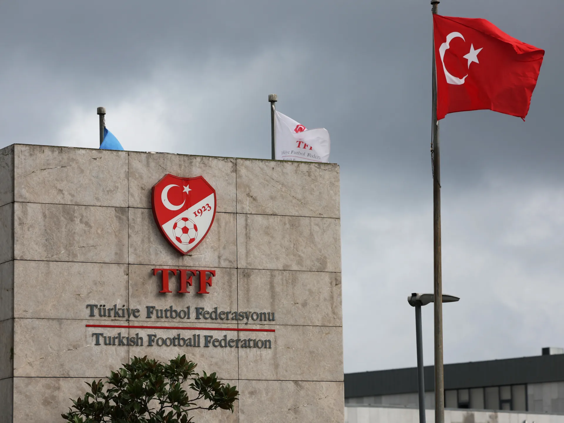 Turkish Football Federation sign and crest beneath a Turkish flag on a cloudy day.