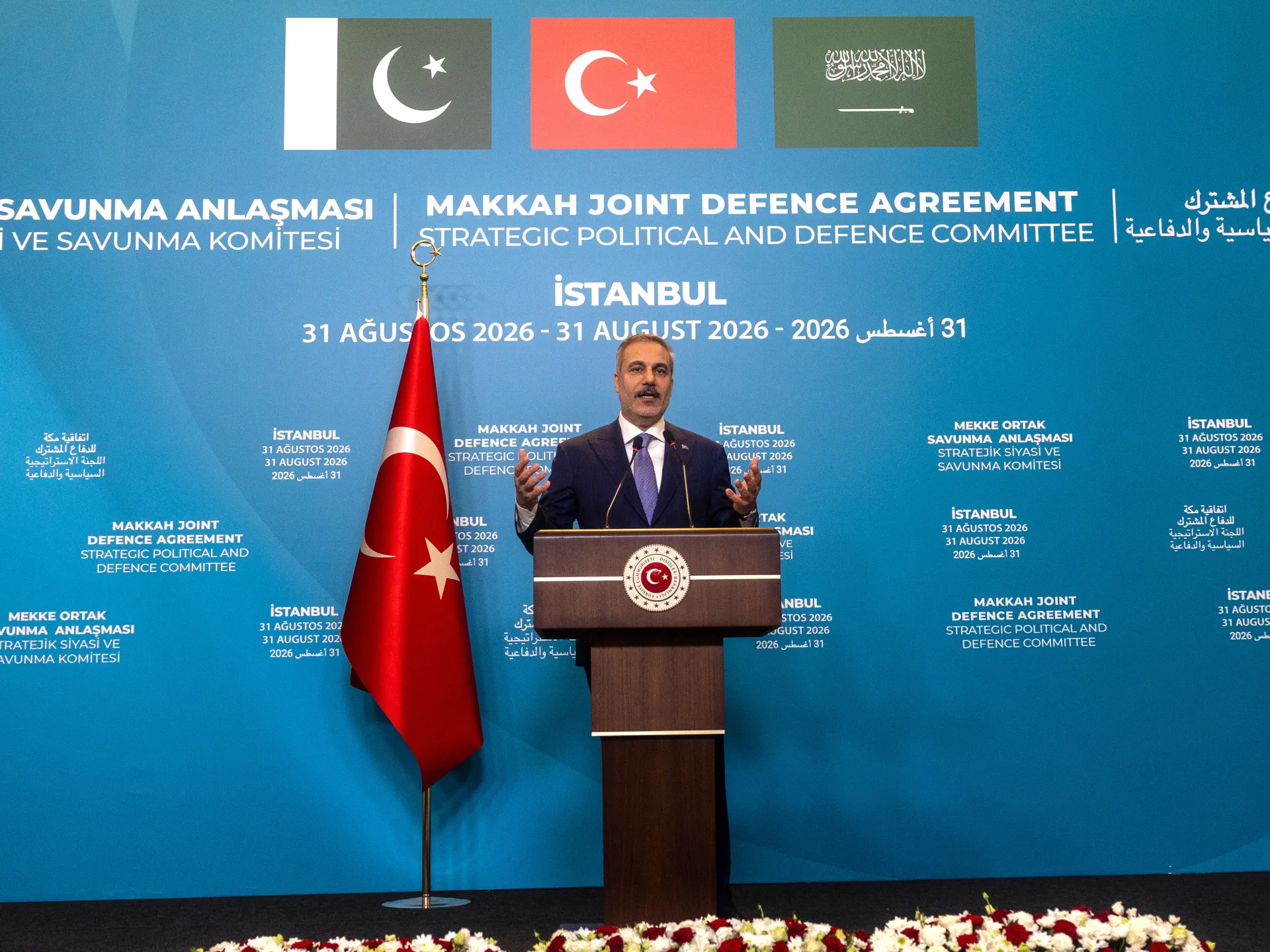 Turkish official speaking at a podium beside a Turkish flag against a blue diplomatic backdrop