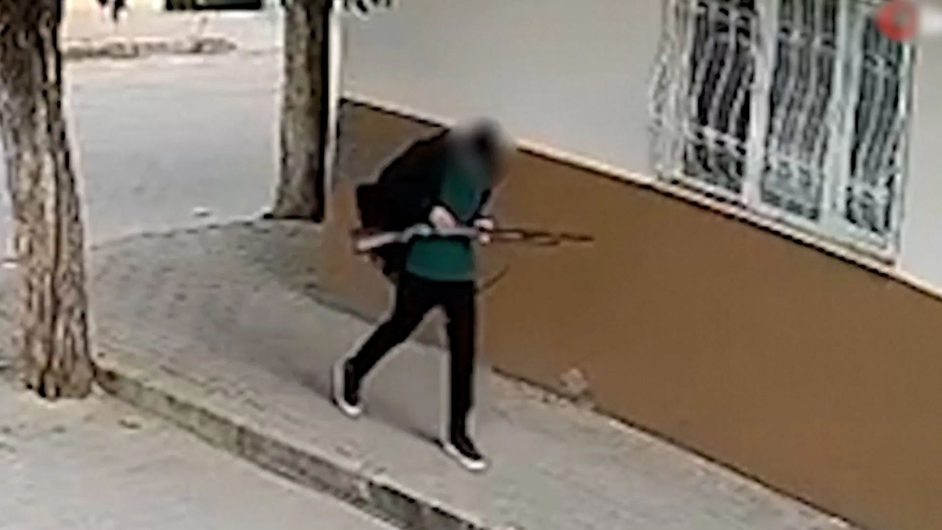 Blurry surveillance image shows a person walking along a sidewalk beside a building.
