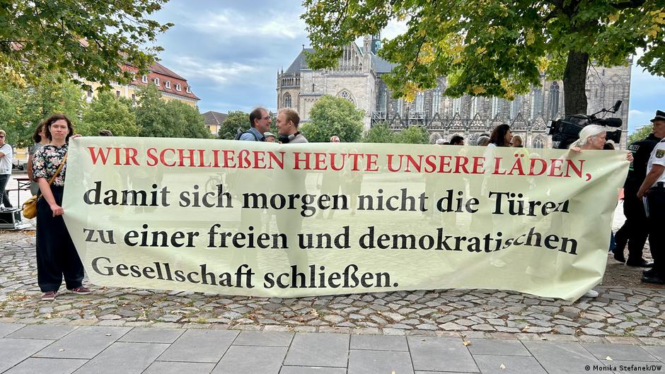 Demonstrators hold a German banner outside a Gothic cathedral, with police and trees behind them.