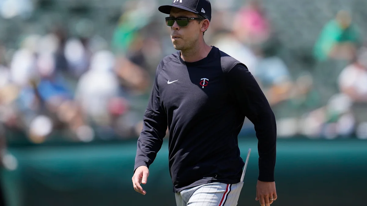 A man in a Twins cap, sunglasses, and black team shirt walks across a baseball field.