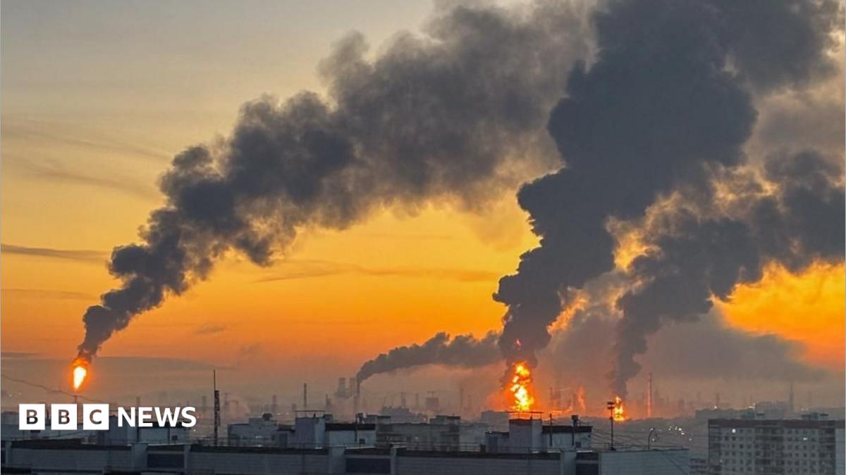 Industrial skyline at sunset with multiple towering smoke plumes and flames rising from distant facilities.