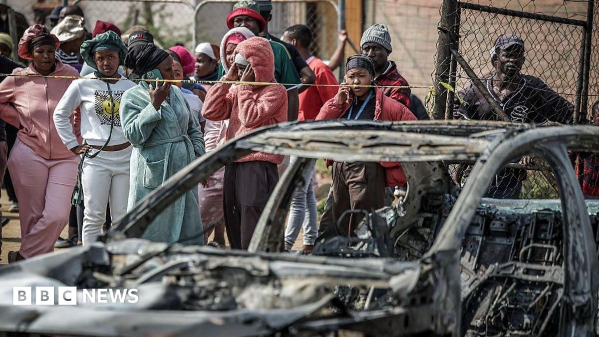 People stand behind a cordon near a charred vehicle, with a wire fence behind them.