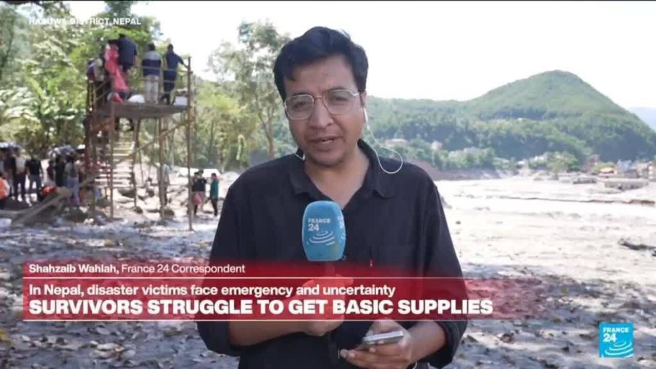 Reporter holds a France 24 microphone beside flood debris, with people and hills visible behind him.