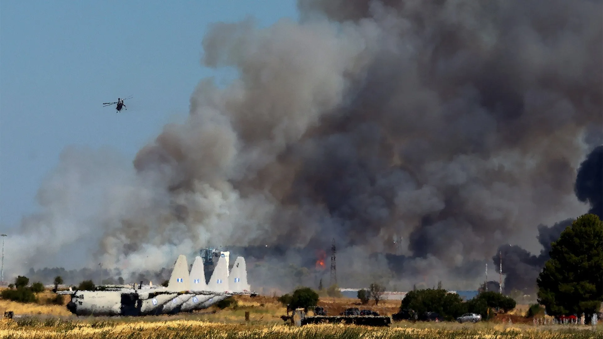 Heavy black smoke rises behind aircraft wreckage as a helicopter flies overhead near a field.