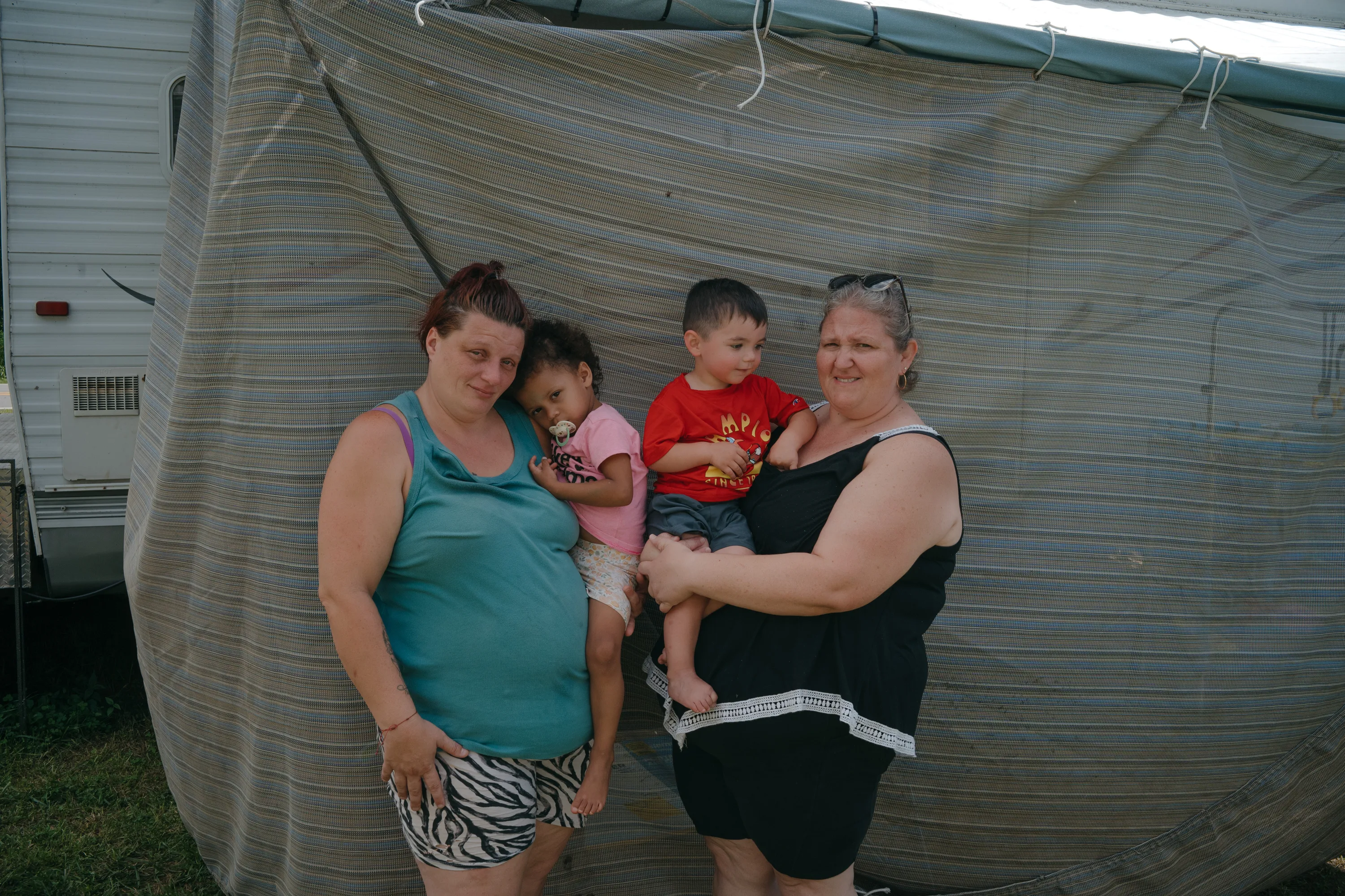 Two women stand with a young girl and boy in front of a striped outdoor canopy.