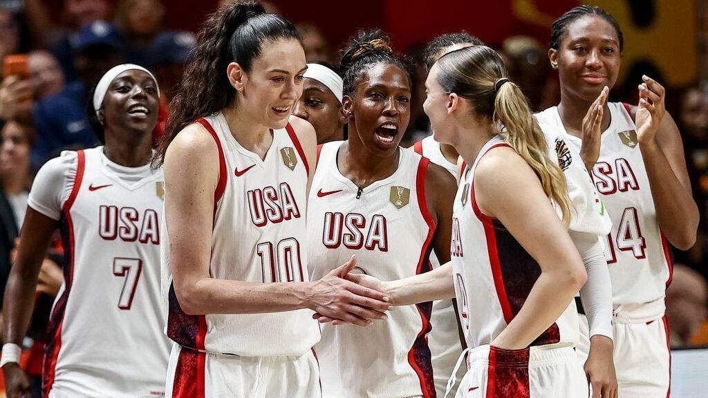 U.S. women's basketball players celebrate together on the court, wearing white USA uniforms.