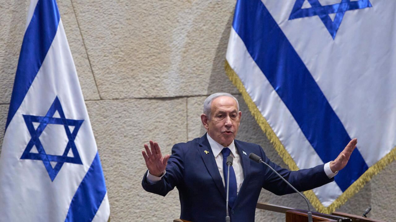 Man in a dark suit speaks at a podium between two Israeli flags.