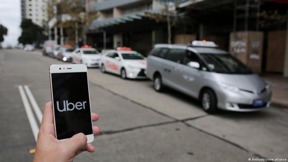 Hand holding a smartphone displaying the Uber logo beside cars with illuminated roof signs
