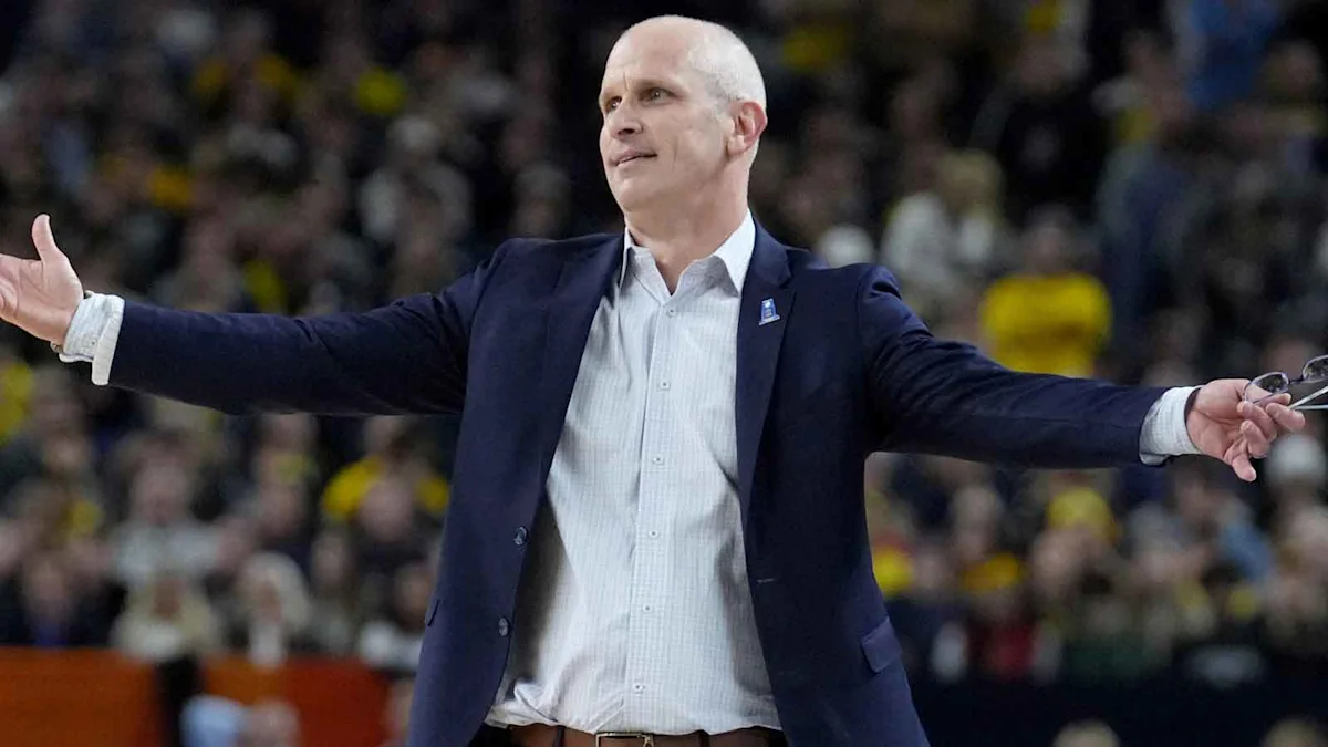 Bald basketball coach in navy blazer stands courtside with arms extended before a blurred crowd