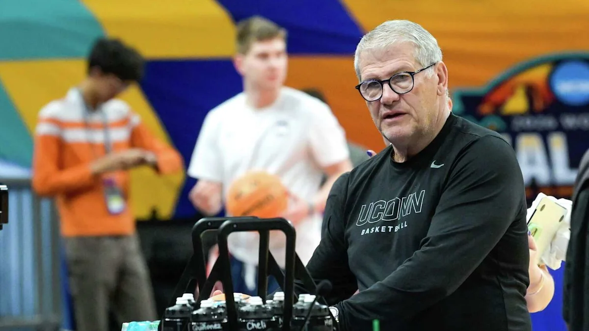 A gray-haired man in glasses wearing a UConn Basketball shirt stands beside equipment at practice.