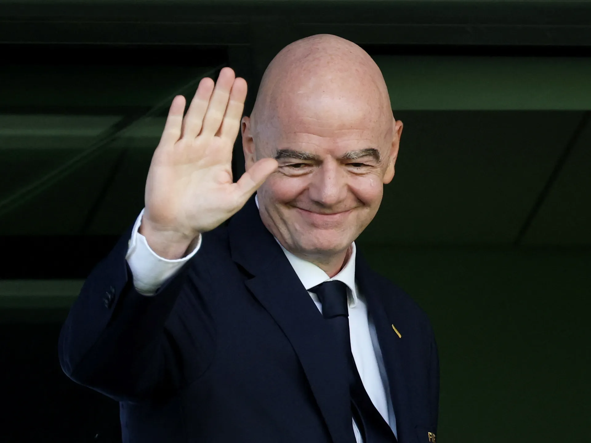 Bald man in dark suit smiling and waving with one hand against a dark background