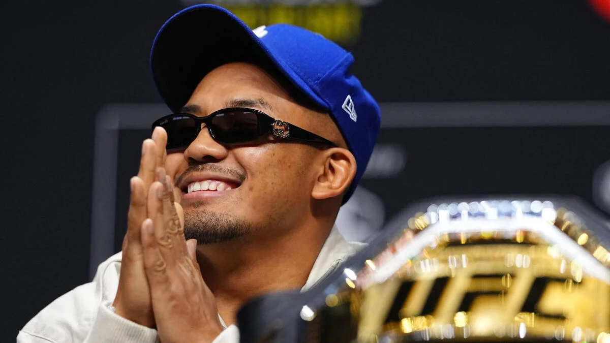 Smiling man in sunglasses and blue cap clasps his hands beside a large gold trophy.