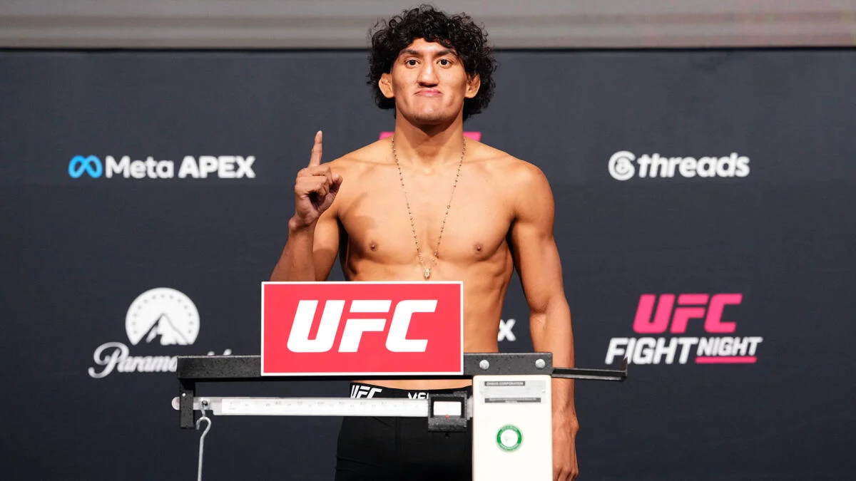 Shirtless fighter raises one finger behind a UFC scale at a weigh-in backdrop.