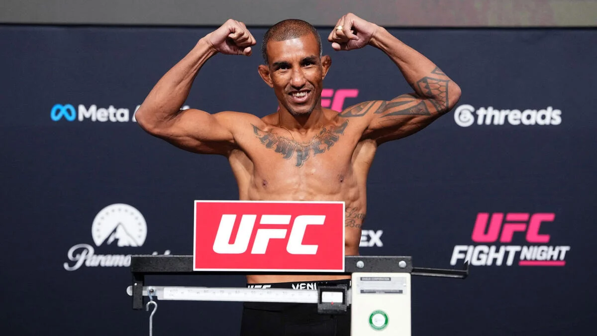 A shirtless fighter flexes at a UFC weigh-in behind a scale bearing a red UFC sign.