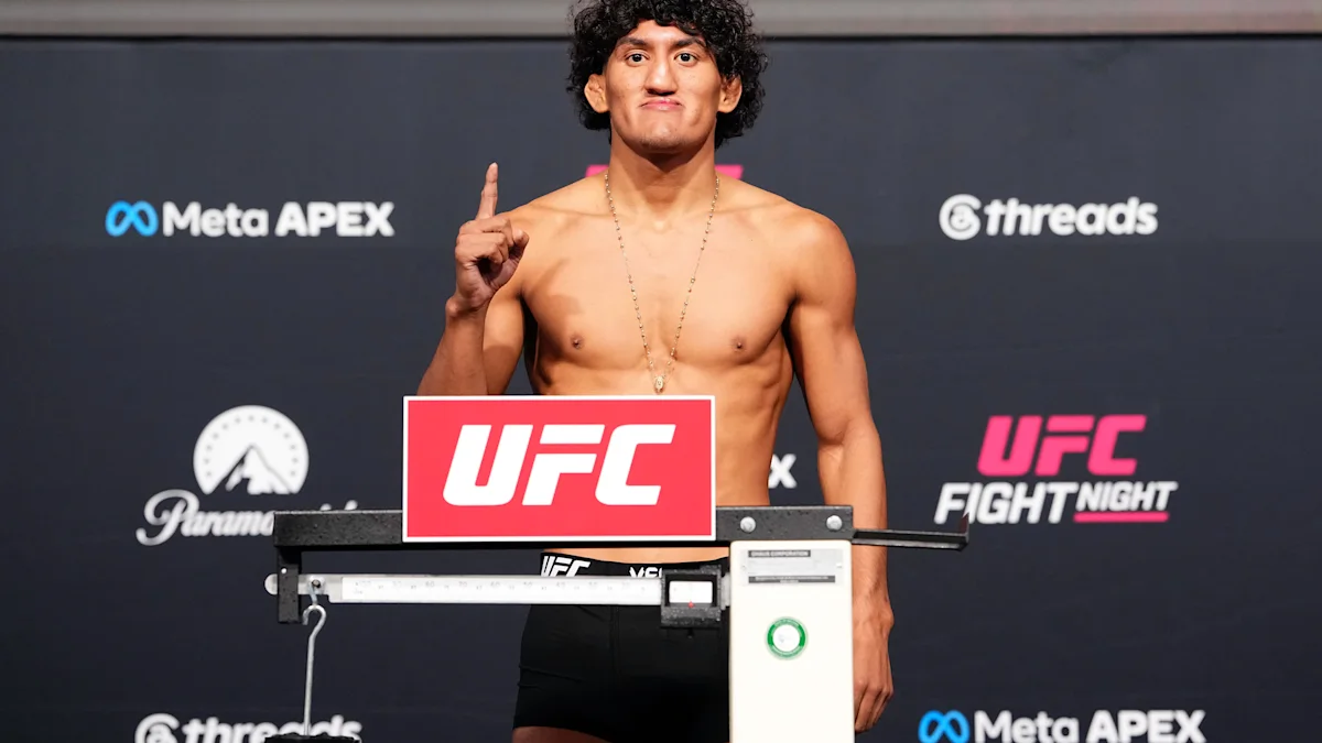 A shirtless fighter raises one finger while standing behind a UFC-branded scale at a weigh-in.