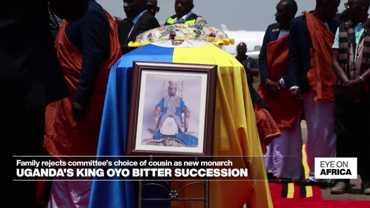 Coffin draped in blue and yellow fabric, floral wreath, framed royal portrait, and mourners gathered outdoors