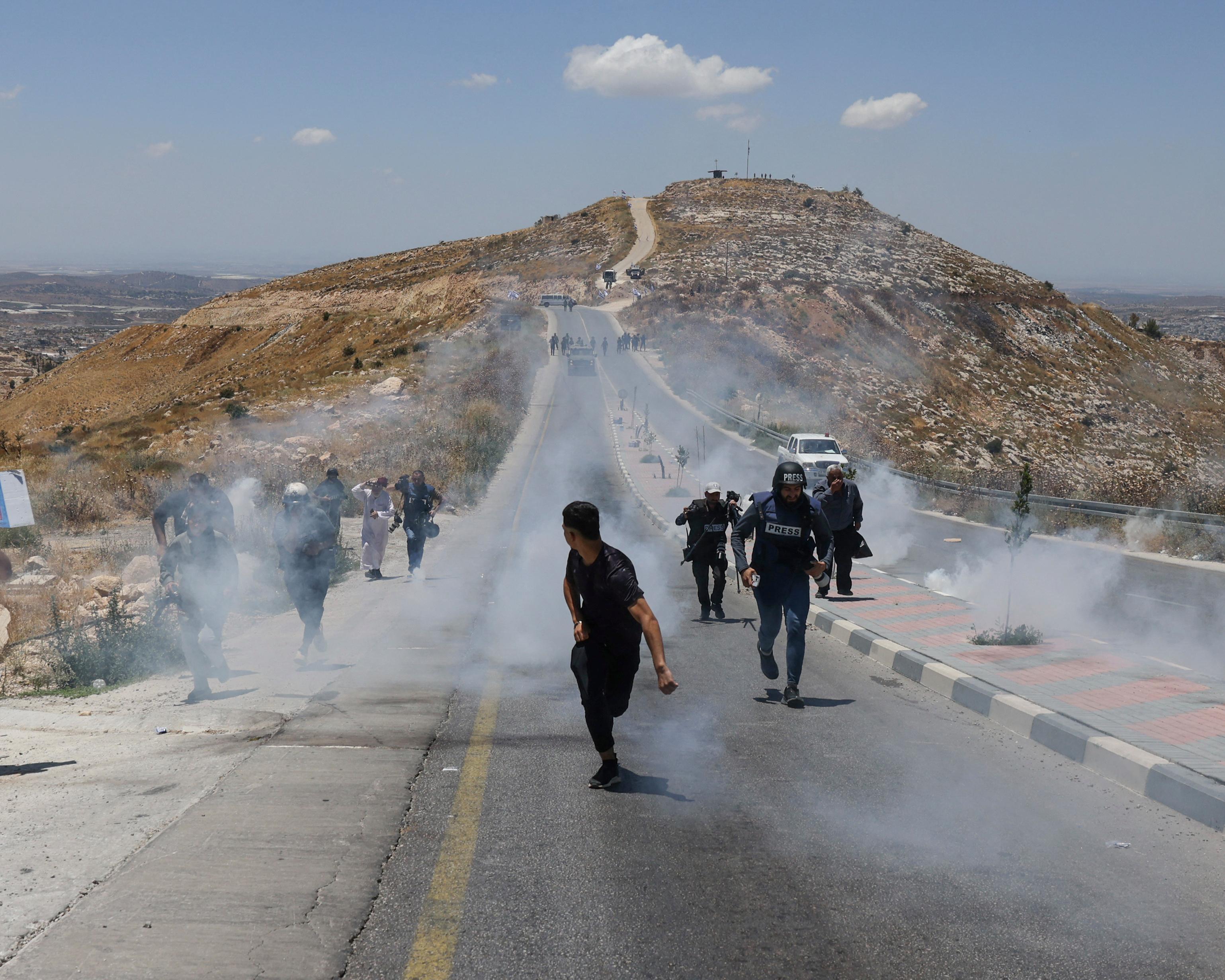 Journalists and other people move through smoke on a road beside a rocky hill