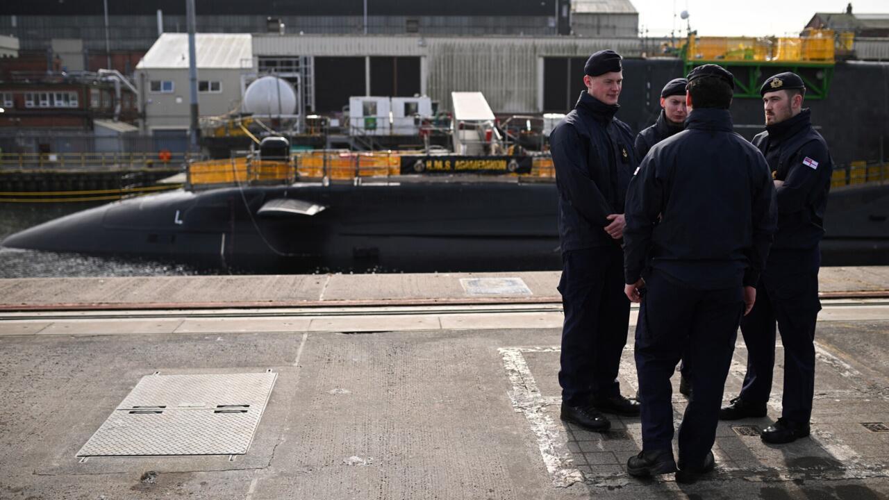 Four naval personnel stand beside a surfaced submarine at an industrial dockyard.