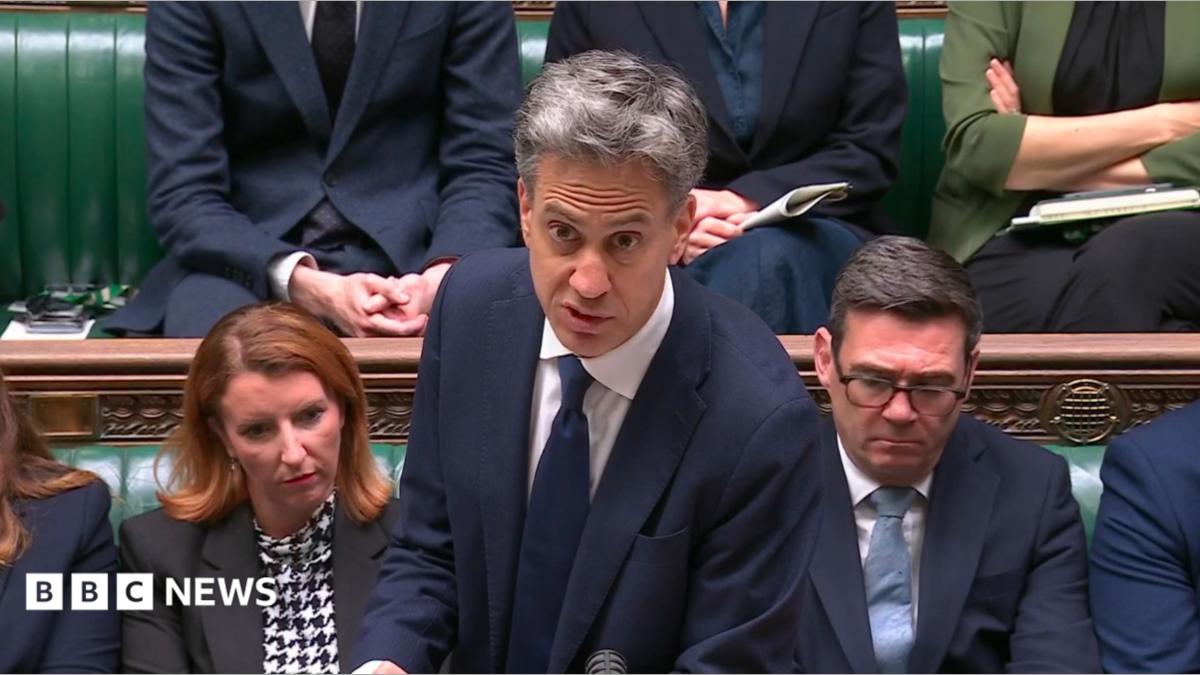 Politician speaking in Parliament with seated colleagues on green benches behind him