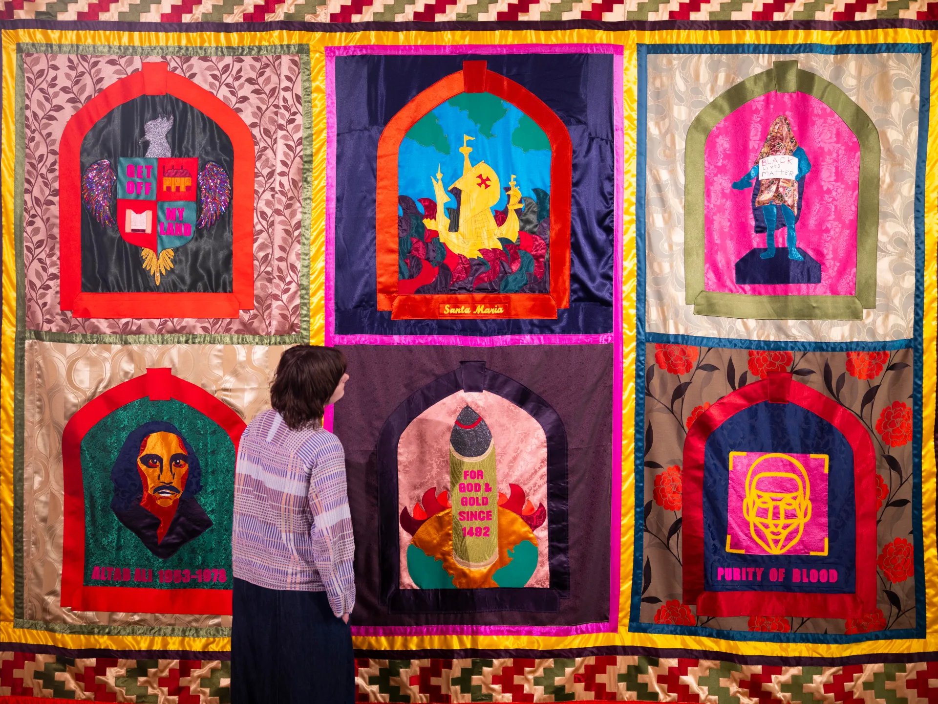 Visitor stands before a colorful textile artwork featuring six arched panels with symbolic imagery.