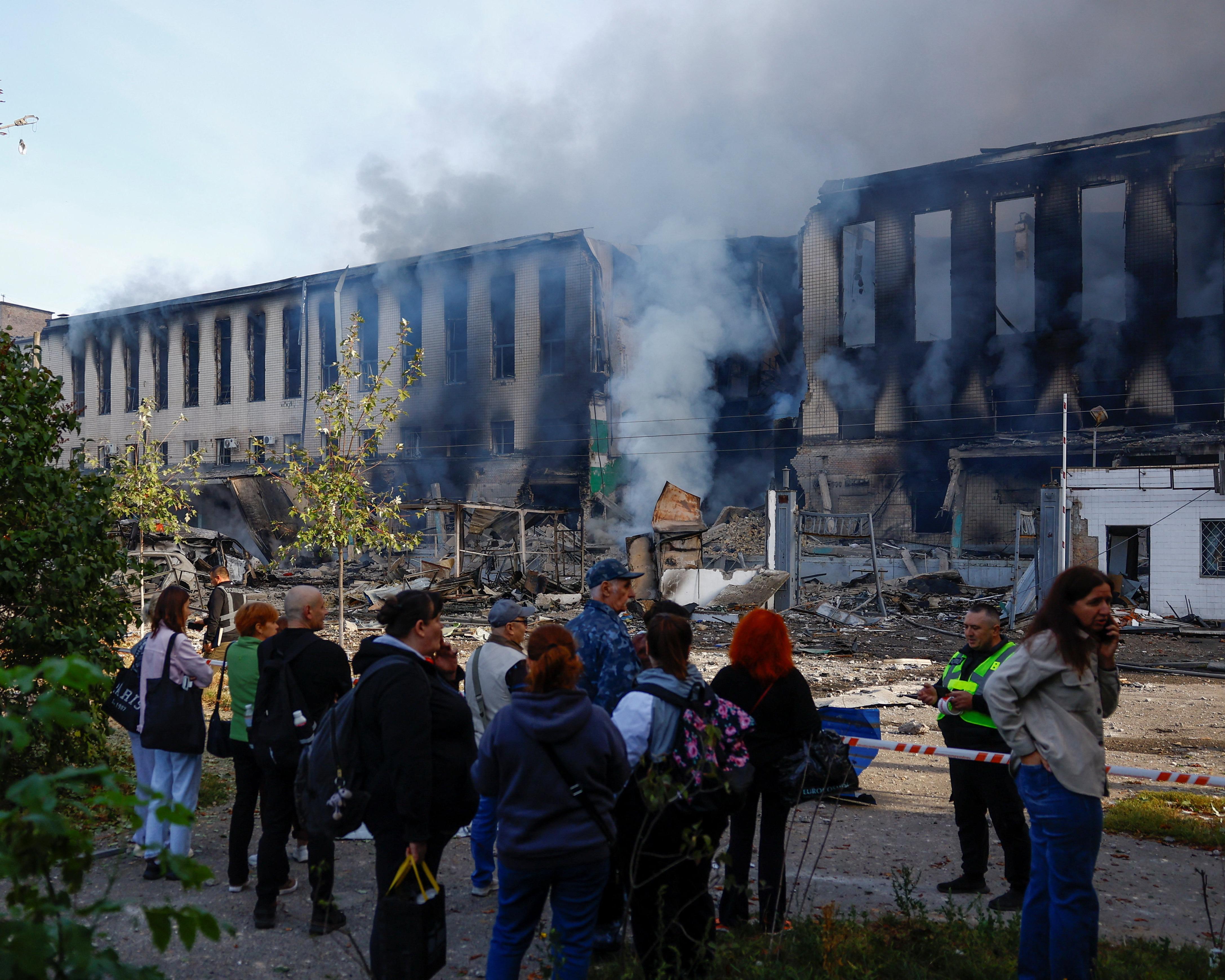 Smoke rises from a heavily damaged building as people gather behind a cordon.