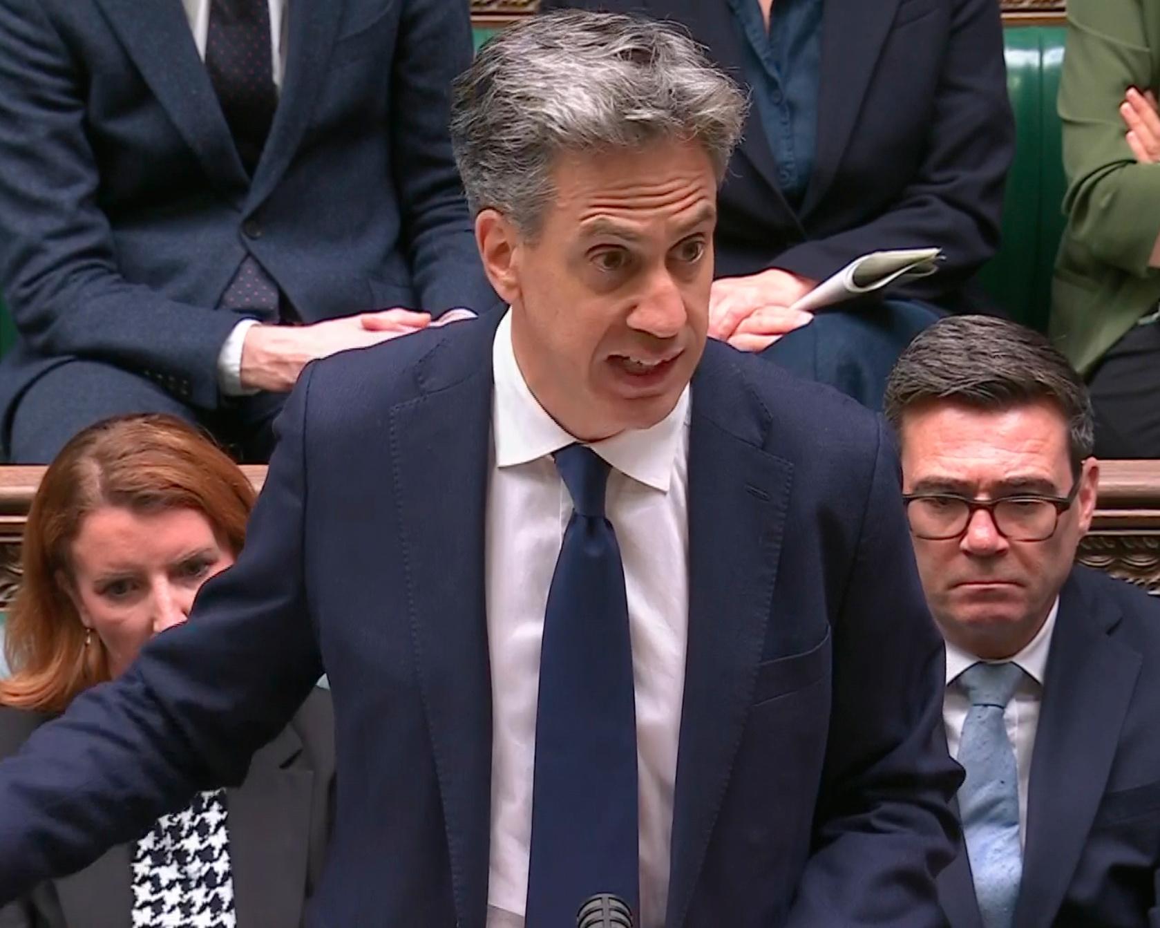 Gray-haired man in a navy suit speaks during a crowded parliamentary session, with seated officials behind him.