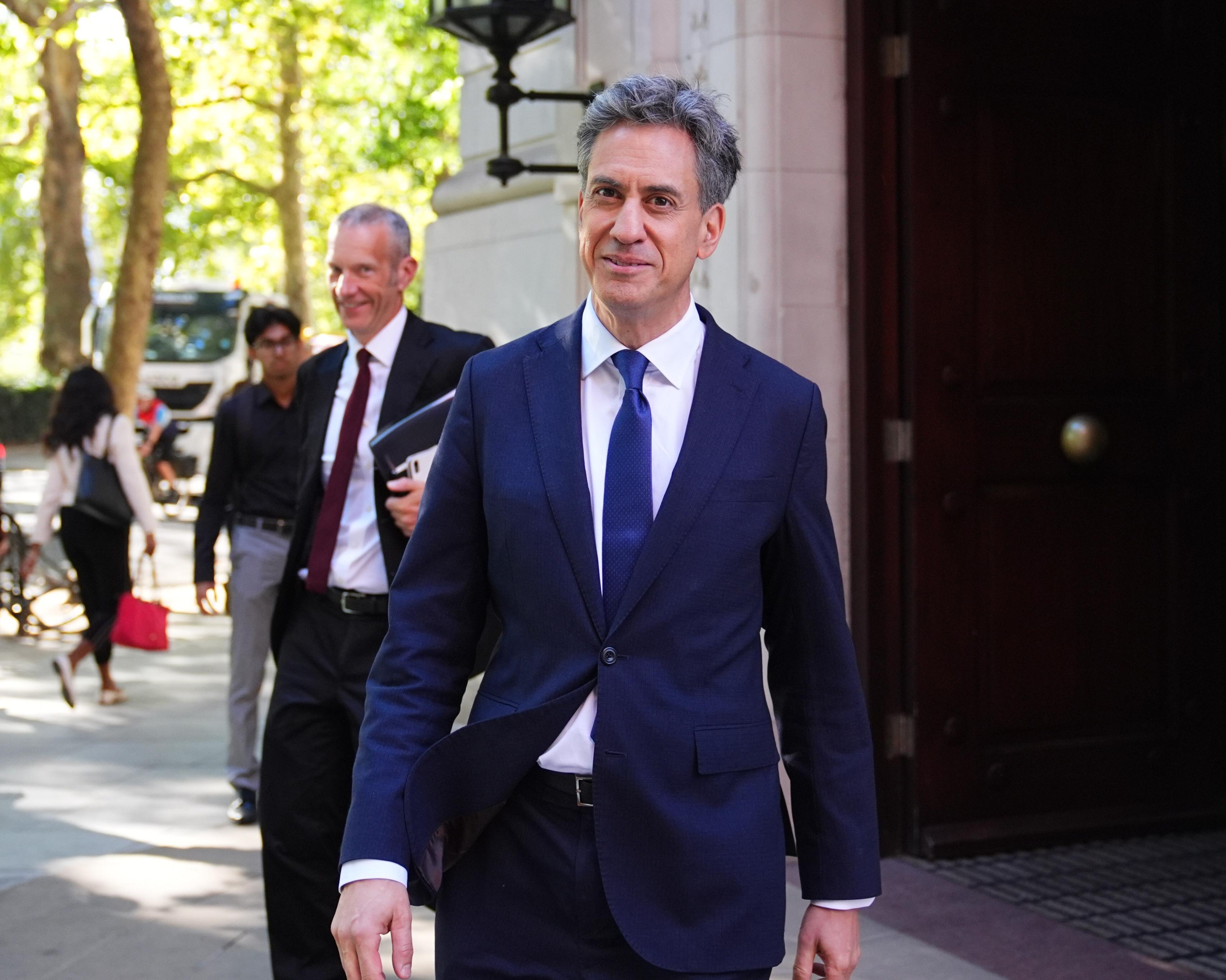 Gray-haired man in navy suit walks outside a building, with colleagues and pedestrians behind him.